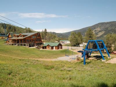 A view of a ski resort base area in summer, featuring a large lodge with a green roof, a smaller wooden cabin, a blue chairlift tower, and a grassy foreground, with forested mountains in the background.
