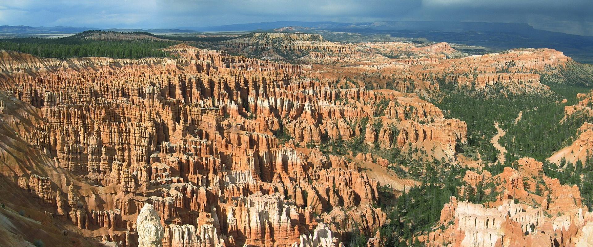 A panoramic landscape photograph showing a vast, deep canyon filled with dense, reddish-orange hoodoo rock formations, with green coniferous trees visible on the rim and in the crevices, set against a cloudy sky.