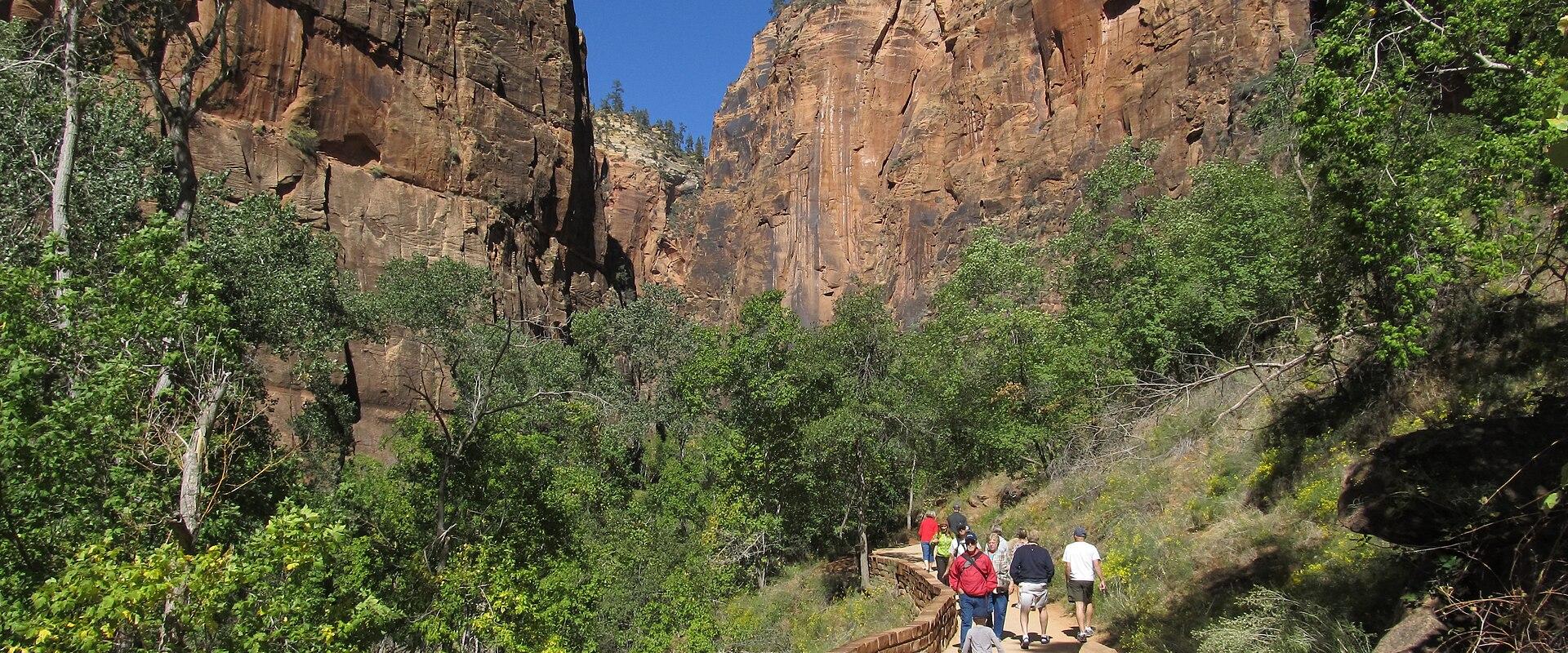 A group of people walking along a paved, multi-level trail with low stone walls, flanked by green trees and vegetation, set against a backdrop of tall, reddish-brown sandstone cliffs.