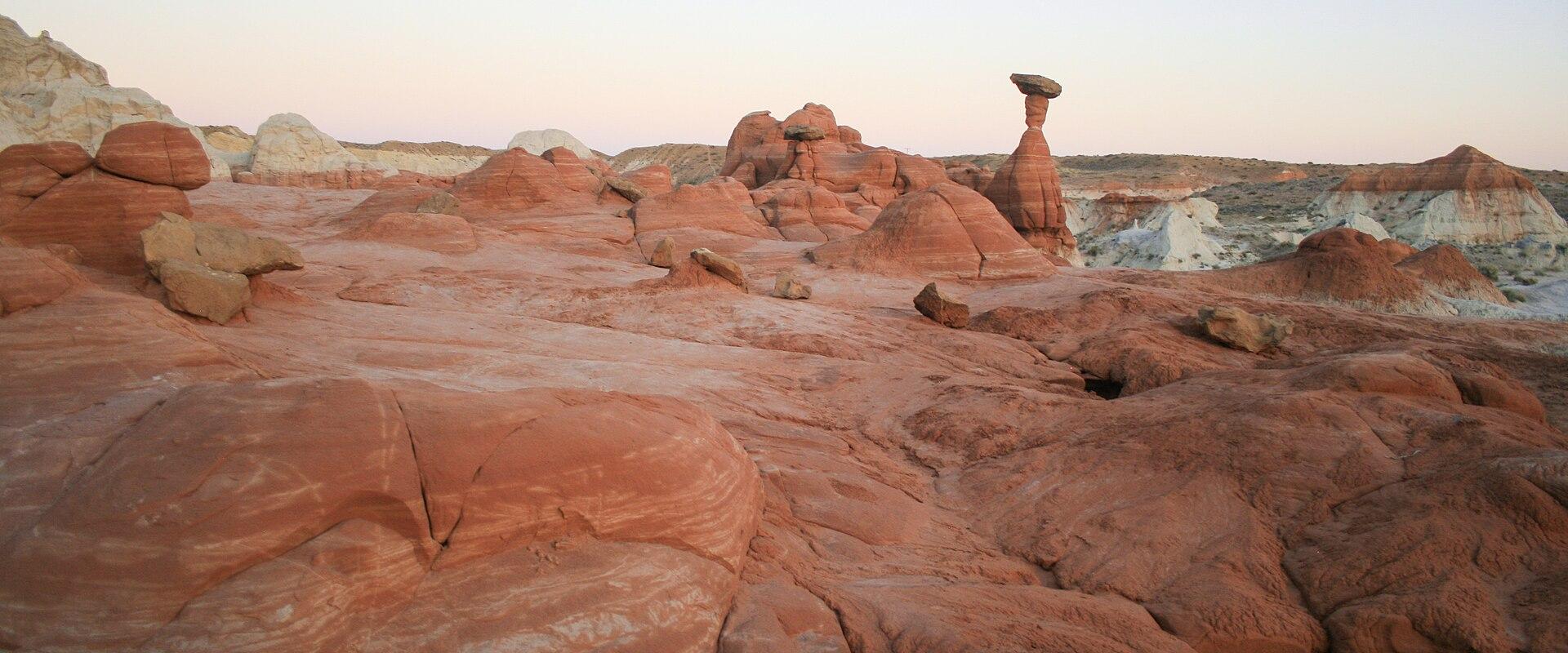 A landscape photograph showing a desert environment with reddish sandstone formations, including a prominent, tall, thin rock pillar (hoodoo) capped by a darker, flat rock, set against a pale sky.