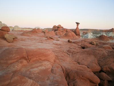 A landscape photograph showing a desert environment with reddish sandstone formations, including a prominent, tall, thin rock pillar (hoodoo) capped by a darker, flat rock, set against a pale sky.