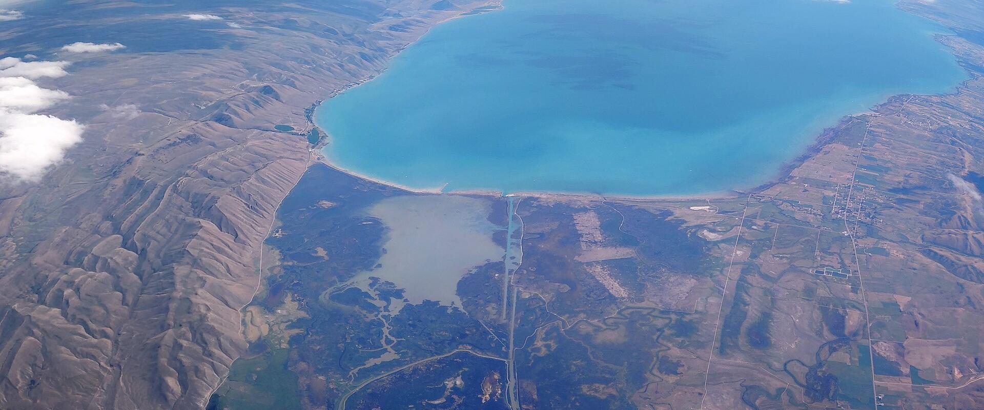 A high-altitude aerial photograph showing a large, turquoise body of water (Bear Lake) bordered by arid, hilly terrain, with a distinct causeway or bridge structure visible in the lower center.