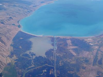 A high-altitude aerial photograph showing a large, turquoise body of water (Bear Lake) bordered by arid, hilly terrain, with a distinct causeway or bridge structure visible in the lower center.