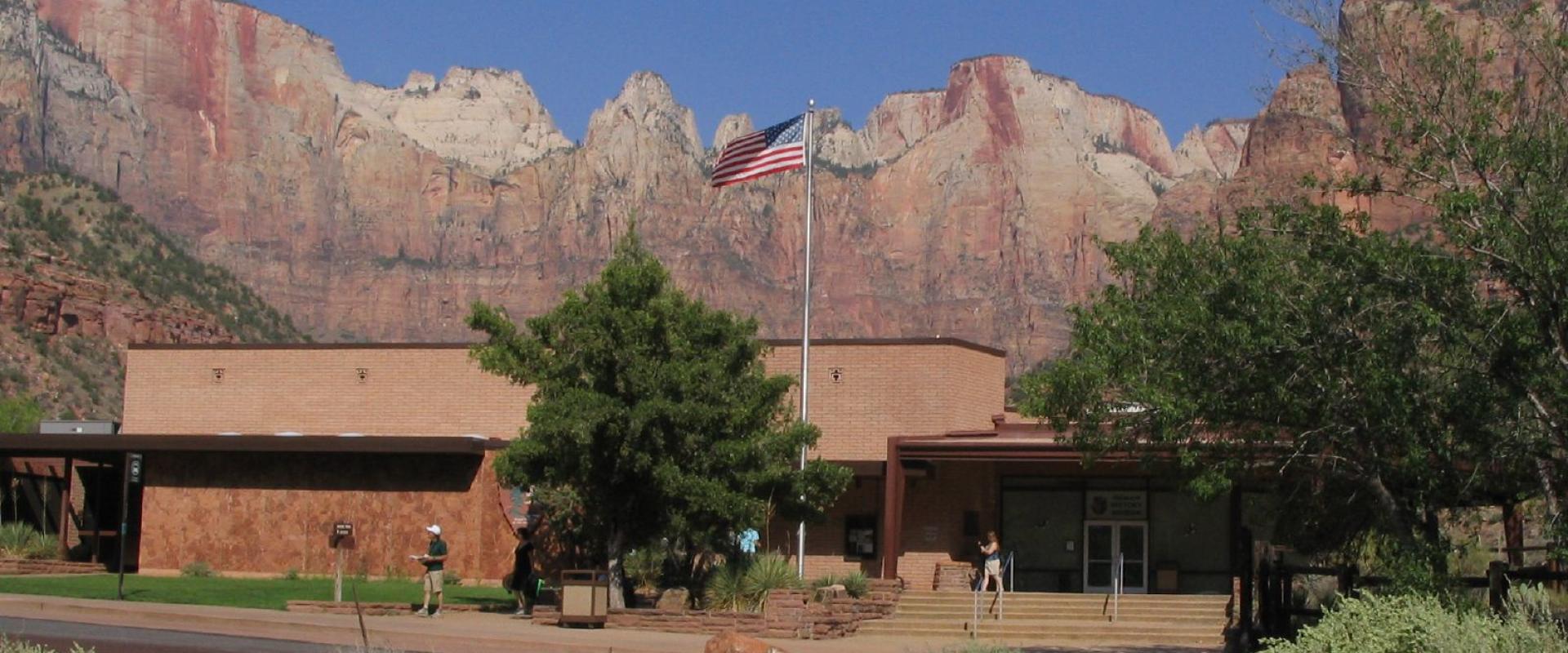 The image shows a low, rectangular building with a flat roof and reddish-brown stucco walls, set against a backdrop of towering red rock cliffs; an American flag flies on a pole in front, and a few people are visible near the entrance.