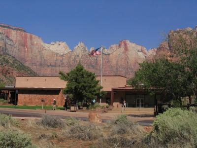 The image shows a low, rectangular building with a flat roof and reddish-brown stucco walls, set against a backdrop of towering red rock cliffs; an American flag flies on a pole in front, and a few people are visible near the entrance.