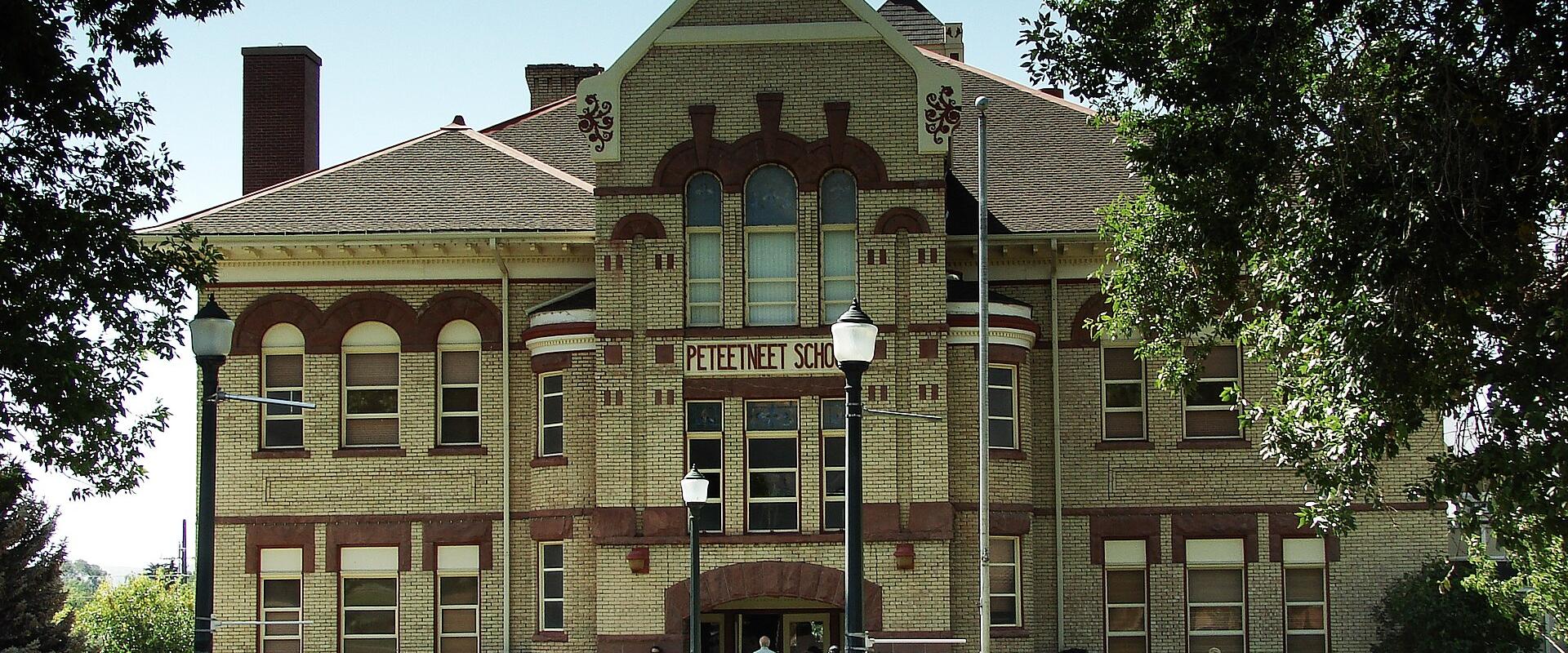 A large, historic brick building with distinctive red brick arches and a gabled facade, featuring the text "PETEETNEET SCHOOL" clearly visible above the main entrance, with people gathered on the lawn in front.