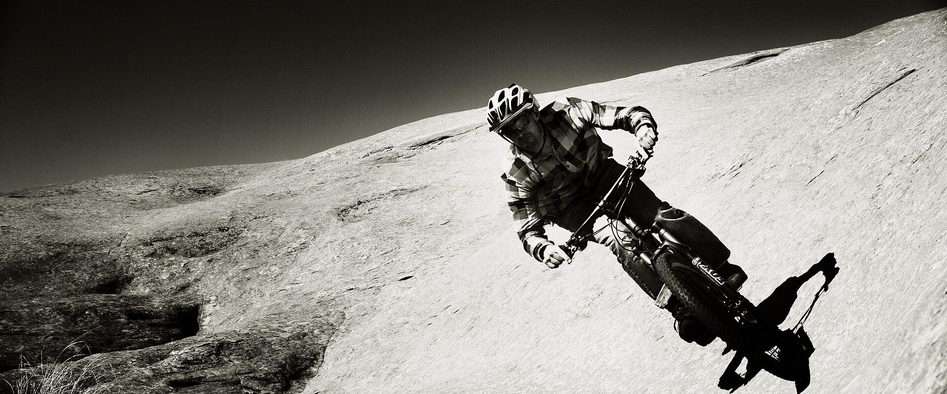 A black and white photograph showing a mountain biker riding down a steep, smooth, sloping rock face, with sparse vegetation visible in the lower left corner.