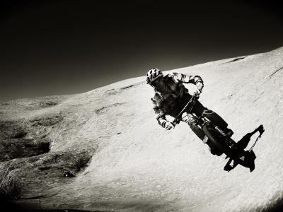 A black and white photograph showing a mountain biker riding down a steep, smooth, sloping rock face, with sparse vegetation visible in the lower left corner.