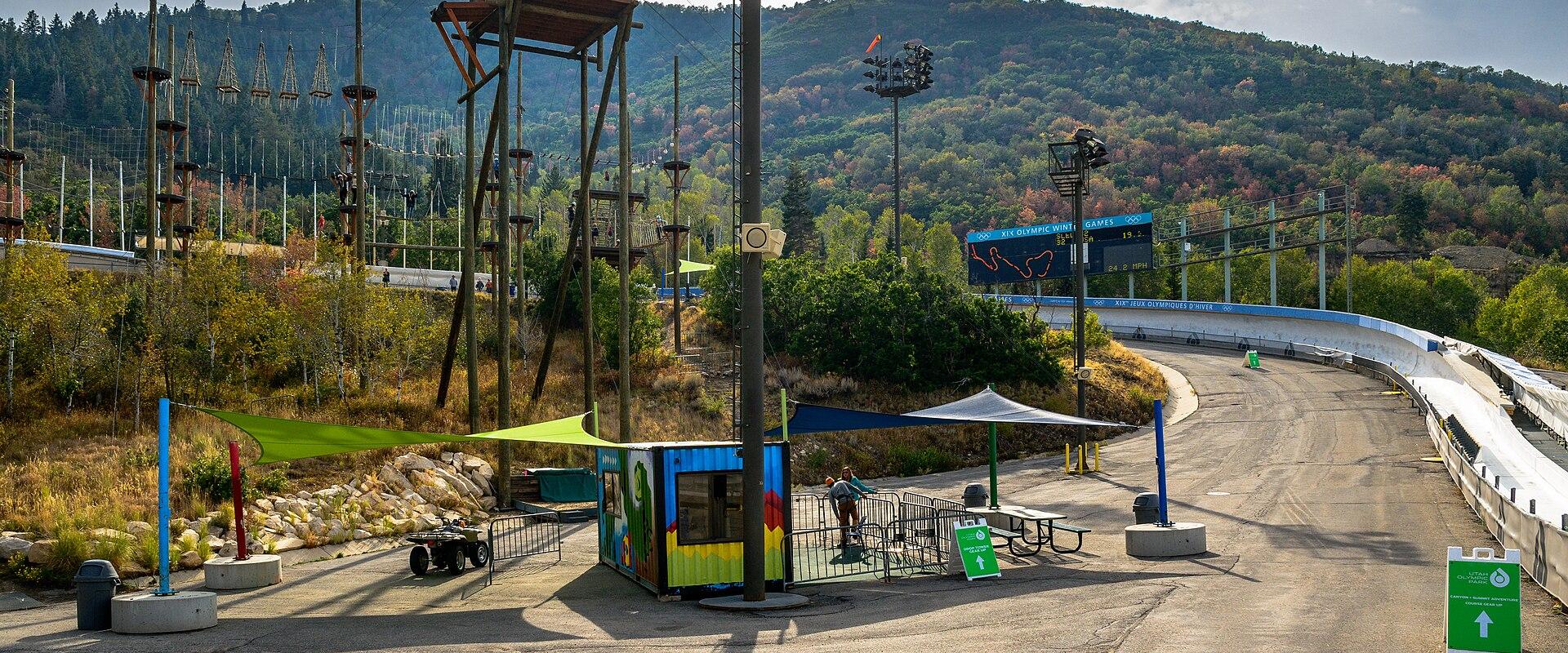The image shows a paved area with a small, colorful booth, a green canopy, and a large, winding track structure (likely a luge or bobsled track) in the background, set against a forested hillside.