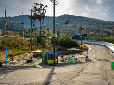 The image shows a paved area with a small, colorful booth, a green canopy, and a large, winding track structure (likely a luge or bobsled track) in the background, set against a forested hillside.