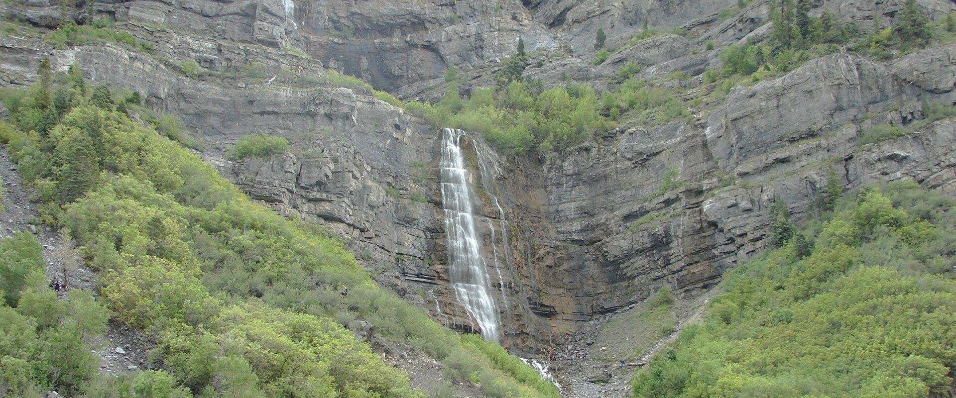A waterfall cascading down a steep, rocky cliff face, surrounded by green trees and vegetation, with a partial view of a structure in the bottom left corner.
