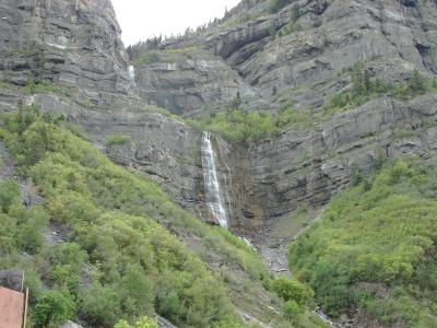 A waterfall cascading down a steep, rocky cliff face, surrounded by green trees and vegetation, with a partial view of a structure in the bottom left corner.
